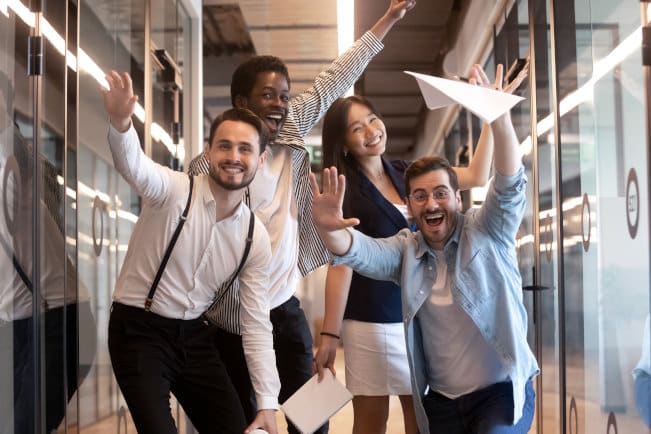 Overjoyed funny multiracial millennial employees look at camera having fun in office hallway, happy excited multiethnic diverse work group, young motivated team posing for picture in corridor