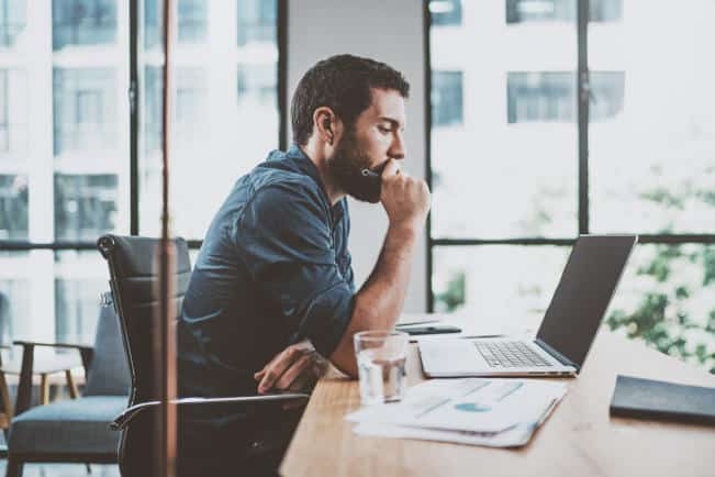Young pensive coworker working at sunny work place loft while sitting at the wooden table.Man analyze document on laptop display.Blurred background.Horizontal